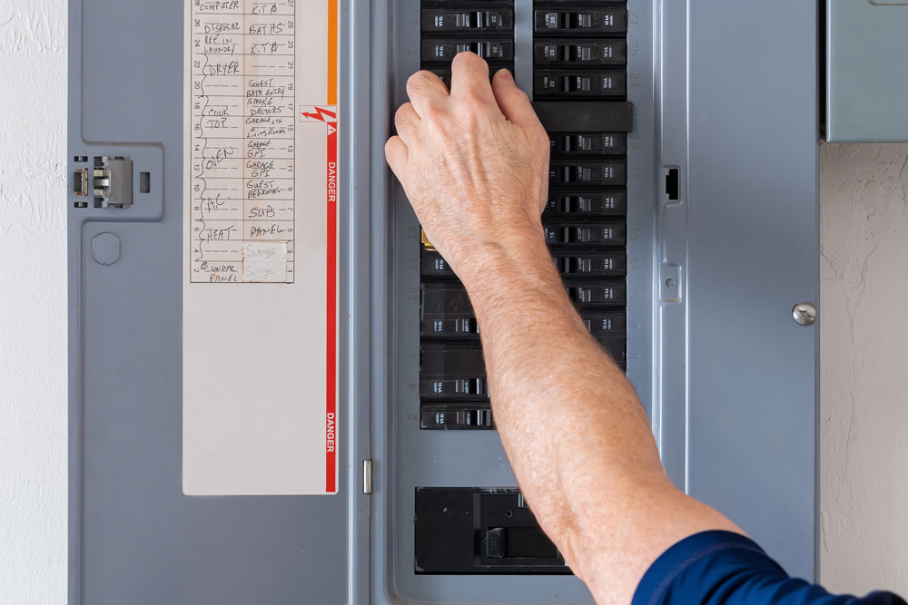 An electrician's arm and hand are visible pulling down a lever within a residential electrical breaker box.
