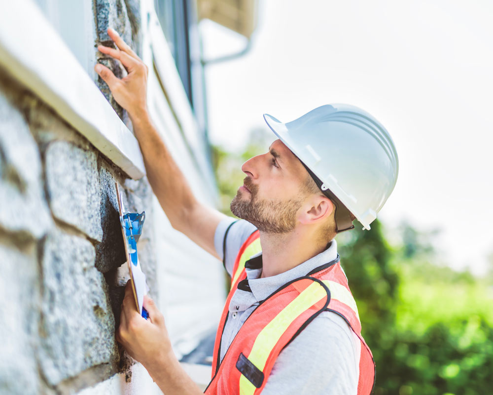 A man in a hard hat and construction vest inspects the outside of a home