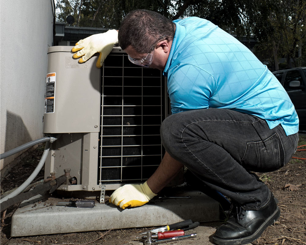 A man kneels down and fixes an HVAC unit outside of a home.