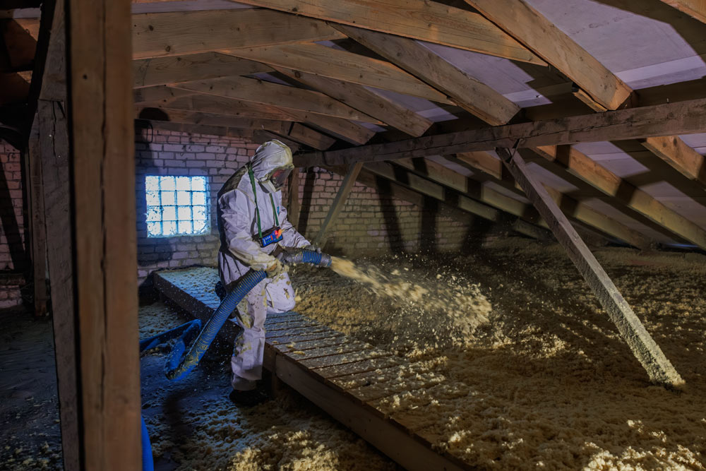 A man in a white hazard suit sprays foam insulation in an attic